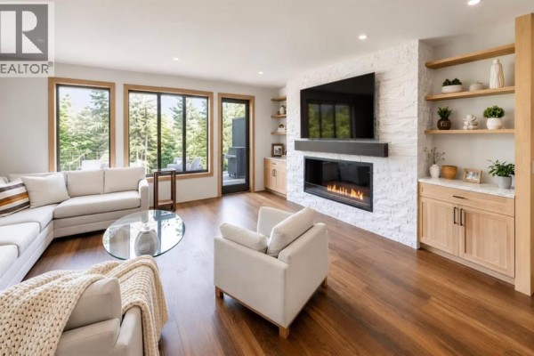 Living room with sectional sofa, armchair, fireplace, and large windows with forest view. - 905a Bridge Road, Canmore