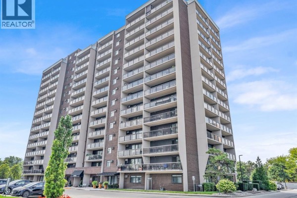 View of a tall brick apartment building with balconies and trees under a blue sky. - 9099 Riverside Unit# 402, Windsor