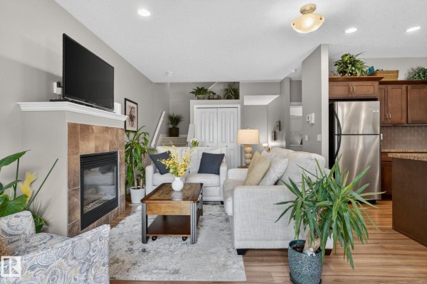 Living room with a fireplace, beige furniture, and plants, next to a small kitchen. - #91 7293 South Terwillegar Dr Nw, Edmonton
