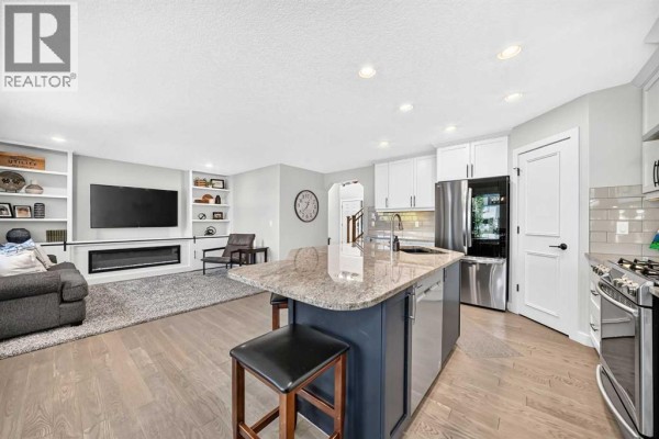 Living room with a large television, fireplace, built-in shelving, and an open view into the kitchen area. - 91 Cranfield Circle Se, Calgary