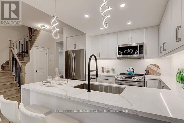 Contemporary kitchen with white island and appliances, adjacent to a staircase and hallway entrance. - 912 - 8110 Birchmount Road, Markham