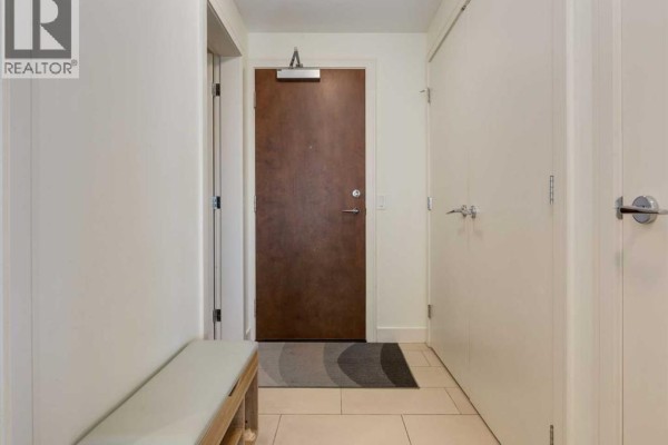 Apartment entryway with a wooden door, beige walls, and a bench with storage. - 913, 222 Riverfront Avenue Sw, Calgary