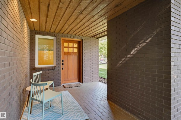 Covered porch entrance with brown brick walls, a wooden door, two chairs, and a patterned rug. - 9130 118 St Nw, Edmonton