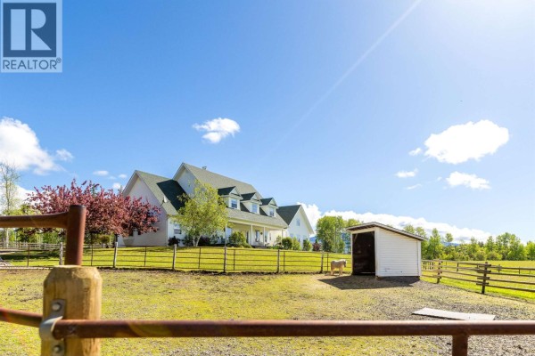 Aerial view of a white house with horse paddocks and surrounding trees under a blue sky. - 9146 Engman Road, Smithers
