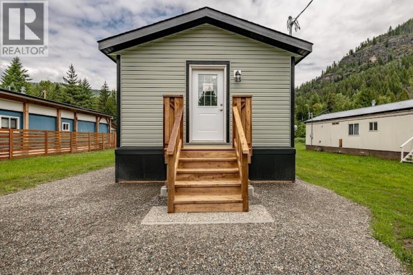 Side view of a grey mobile home with two entrances and wooden steps, surrounded by grass. - 916 Airport Road Unit# 7, Salmo