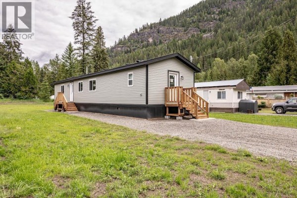Longer side view of the grey mobile home showing a gravel driveway, truck, and wooded mountains. - 916 Airport Road Unit# 7, Salmo