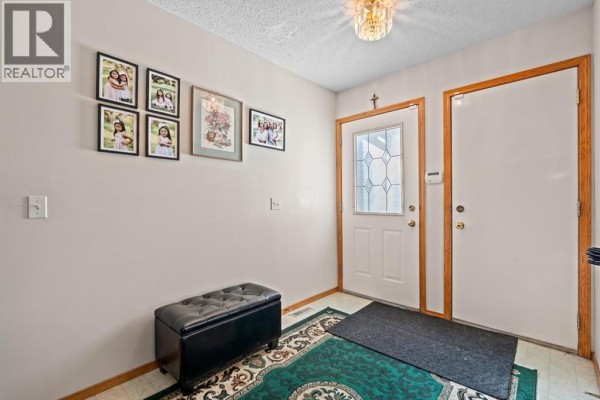The entryway has light beige walls, a black tufted storage bench, and a green patterned rug. - 92 Del Ray Close Ne, Calgary