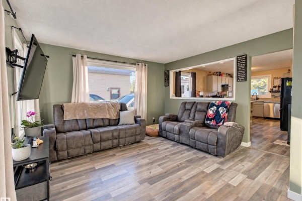 Living room with grey reclining sofas, light wood laminate flooring, and an open view into the kitchen. - 9218 81a St, Fort Saskatchewan