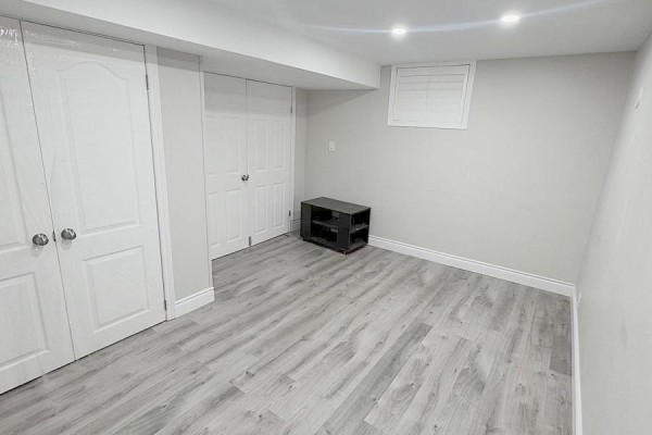 A finished basement room with light wood-look vinyl flooring, white bi-fold closet doors, and a black media console. - 925 Byron Street N, Whitby