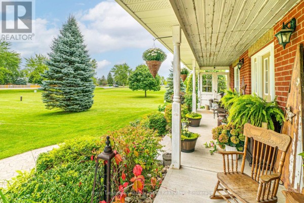 View from the covered porch showing landscaping, a rocking chair, and a large green lawn. - 929 Eldon Road, Kawartha Lakes