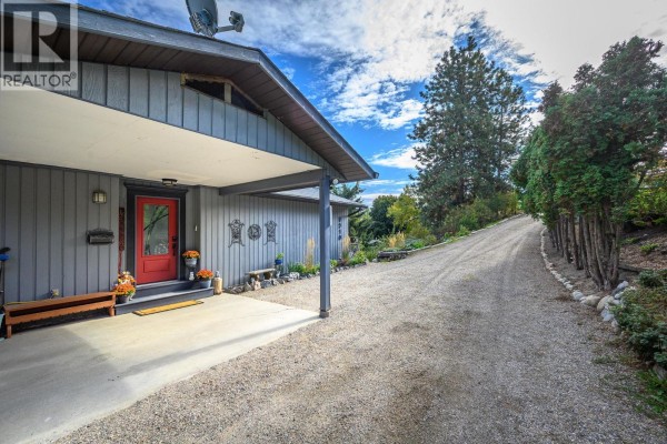 Driveway curving up to the grey home, past tall evergreen trees and showing the front entrance. - 9310 Northcott Drive, Coldstream