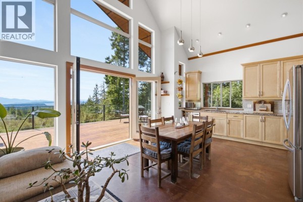 Dining area with wooden table, chairs, and kitchen cabinets next to large windows with forest view. - 9340 Stephens Way, Halfmoon Bay