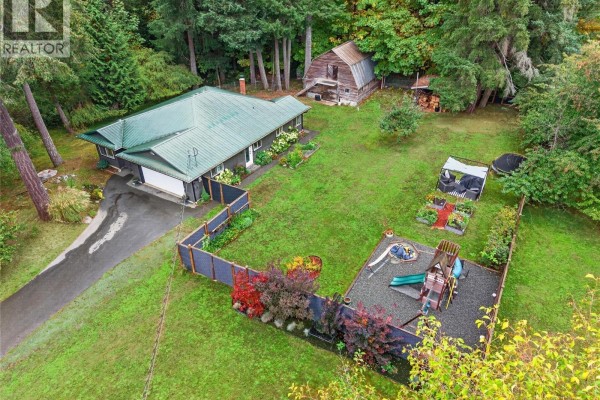 Aerial view of a house with a fenced yard, including a playground and garden beds, next to a wooded area. - 936 Arden Rd, Courtenay