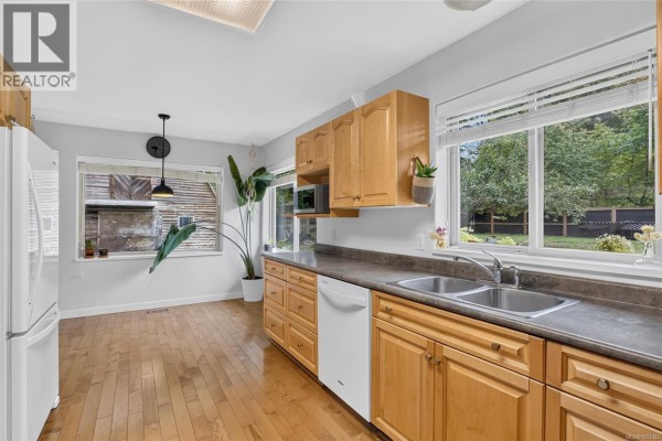 A bright kitchen with light wood cabinets, laminate countertops, a double sink, and large windows overlooking the backyard. - 936 Arden Rd, Courtenay