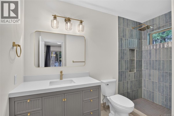 A modern bathroom with a grey vanity, white countertop, gold fixtures, and a walk-in shower with blue tiled walls. - 936 Arden Rd, Courtenay