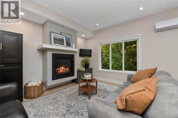 Living room with gas fireplace, grey sofa, and large window with green treed view. - 937 Echo Valley Pl, Langford