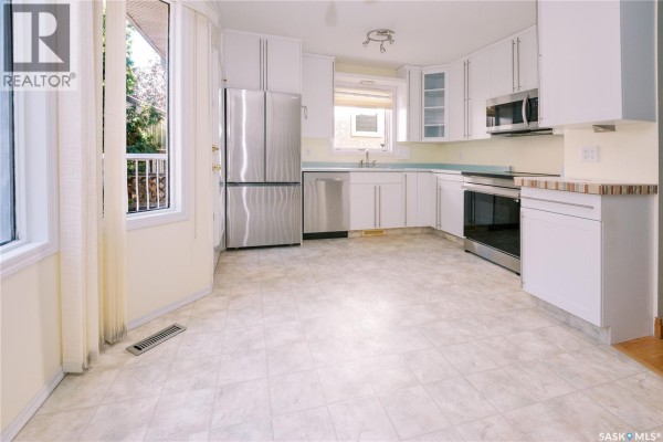 Wooden dining table with four chairs in a sunlit kitchen area with large windows. - 939 Haslam Crescent, Saskatoon