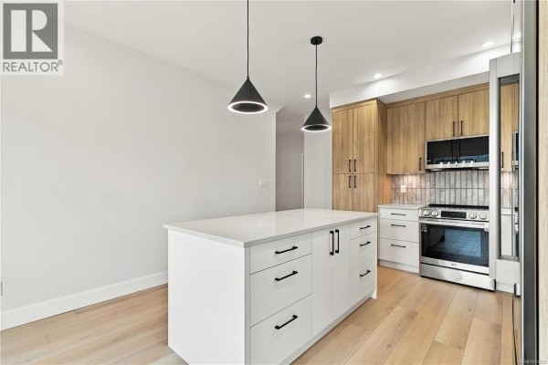 Island kitchen with white cabinetry, light wood cabinets, stainless steel appliances, and two modern pendant lights. - 941 Malbec Link, Langford