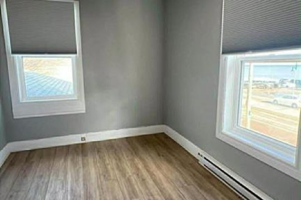 Empty room with grey walls, light wood laminate flooring, and two windows with grey blinds showing an exterior view. - 9418 Main, Richibucto