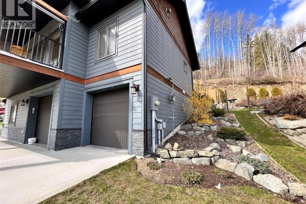 Side exterior of grey house with two garage doors and landscaped rocky slope. - 944 Redstone Drive, Rossland