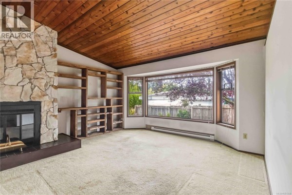Living room with a stone fireplace, built-in wood shelving, bay windows, and a wood plank ceiling. - 946 Paconla Pl, Central Saanich