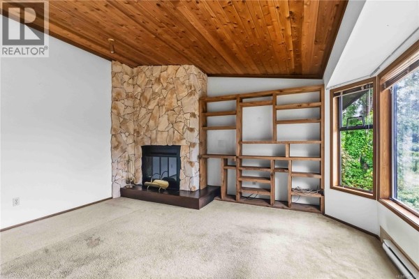 Living room fireplace with stone surround and wood shelving unit; view of trees outside window. - 946 Paconla Pl, Central Saanich