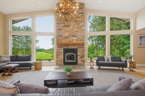Living room with a large stone fireplace, expansive windows overlooking a green field, and modern gray sofas. - 9472 222 Street, Langley