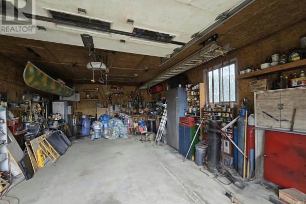 Interior view of a spacious garage filled with tools, equipment, storage shelves, and a canoe suspended. - 954 Cashin Crescent, Labrador City