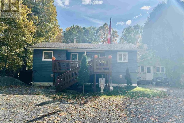 Front view of a blue house with a wooden deck and stairs, surrounded by trees and gravel driveway. - 96 Olar Side Rd, Goulais River