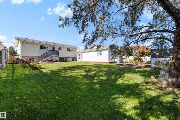 Backyard view of a house with a large lawn, mature tree, wooden deck, and a detached garage. - 9703 104 St, Morinville