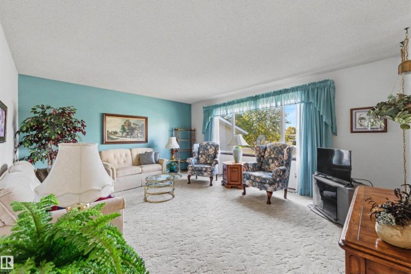 Living room with a textured beige carpet, blue walls, floral armchairs, a sofa, and a large window. - 9703 104 St, Morinville