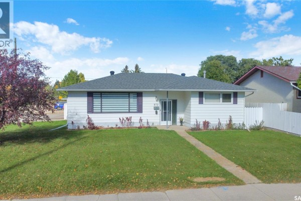 Side exterior view of the white bungalow with a green lawn and mature trees. - 972 Simcoe Street, Moose Jaw