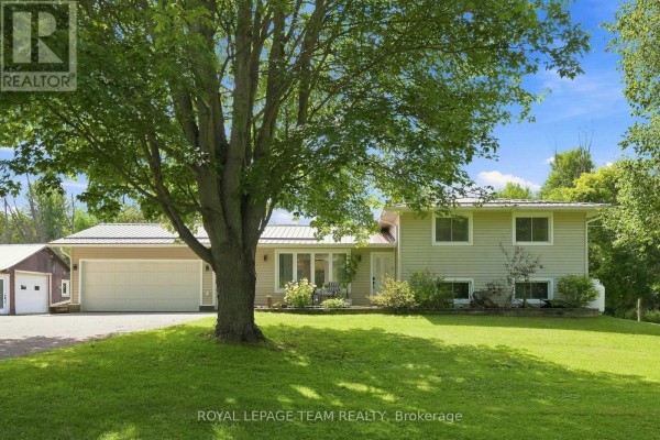Front view of a beige house with a metal roof, green lawn, and mature trees. - 980 County Rd 44, North Grenville