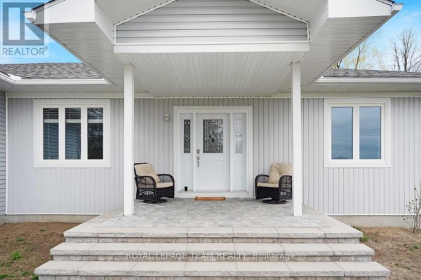 Close-up of the front entrance of a light gray house with a covered porch and two chairs. - 986 South Gower Drive, North Grenville