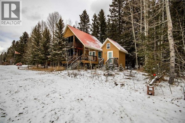 Front view of a wooden cabin with a red metal roof and a small detached outbuilding surrounded by trees and snow. - 99, 5227 Township Road 320, Rural Mountain View County