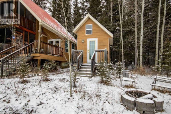 Close-up of a small wooden outbuilding with a white door and steps, next to a larger cabin surrounded by trees and snow. - 99, 5227 Township Road 320, Rural Mountain View County