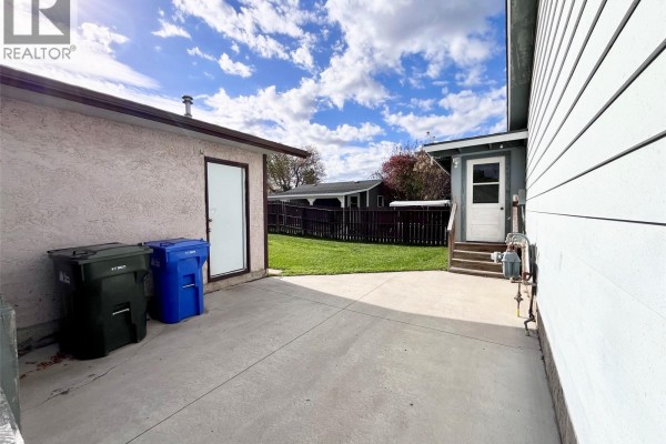 Concrete backyard patio area with two large garbage bins and a door leading to the house. - 99 Bowerman Crescent, Prince Albert