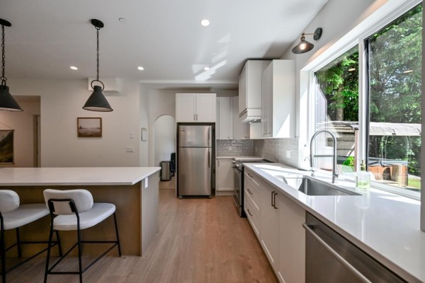 A two-story white house surrounded by dense green trees, with a gravel driveway and a garden. - 9933 Watt Street, Mission