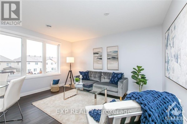 Living room with large windows, grey velvet sofa, glass coffee table, and white armchair. - B - 444 Via Verona Avenue, Ottawa