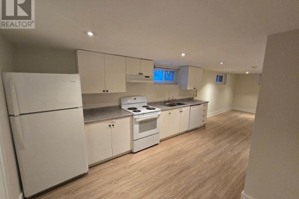 Wide view of a basement apartment kitchen with white appliances, cabinetry, and light wood laminate flooring. - B - 50 Elmer Avenue, Orillia