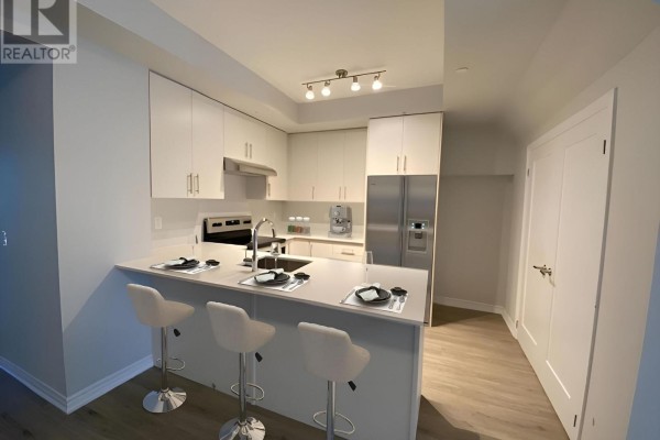 Modern white kitchen with island, bar stools, stainless steel refrigerator, and light wood floors. - B-208 - 349 Wheat Boom Drive, Oakville