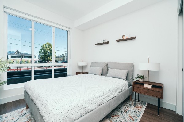 Bedroom with a large window overlooking the street, a grey bed with white patterned bedding, and two bedside tables. - B11 45815 Princess Avenue, Chilliwack Downtown, Chilliwack