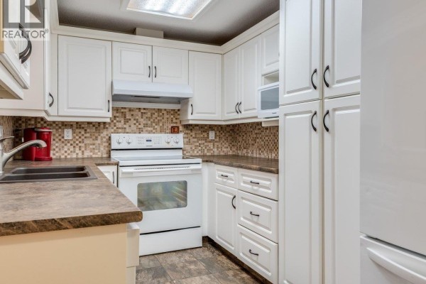 Close-up view of a white kitchen with cabinets, a stove, and a double sink. - B207 4821 53 Street, Ladner
