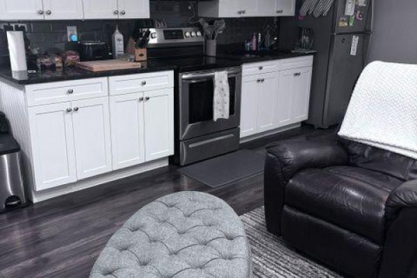 A basement living area with a dark recliner, tufted ottoman, and a galley kitchen in the background. - Basement - 2544 Pillette Road, Windsor