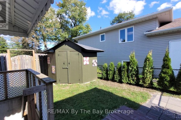 Backyard view of a green shed, manicured trees, and a chain-link fence with a Canadian flag. - Basement - 48 Caribou Trail, Wasaga Beach