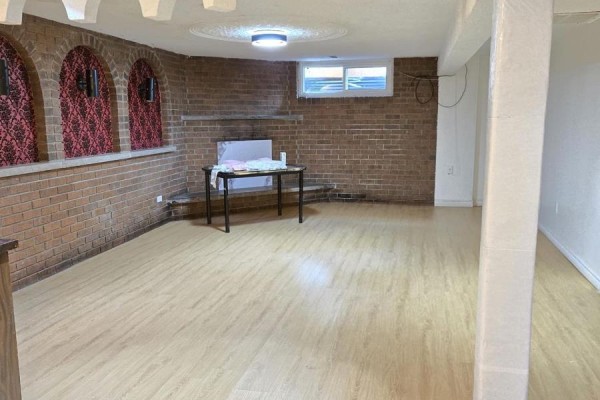 Basement recreation room with wood-look flooring, brick wall, fireplace, and window. - Basement - 61 Hardwick Court, Toronto