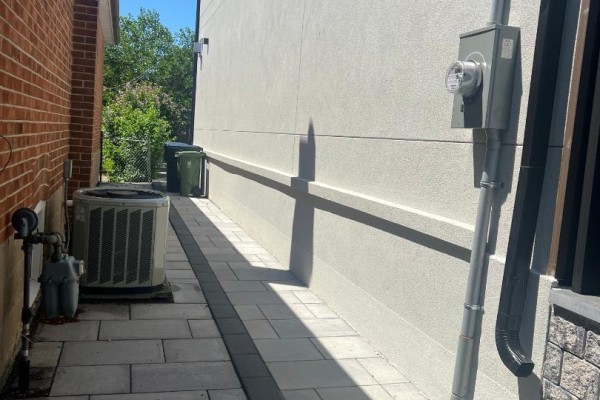 Side walkway with interlocking pavers, a beige stucco wall, and an outdoor electrical meter. - Basement Apartment - 50 Brawley Avenue, Toronto