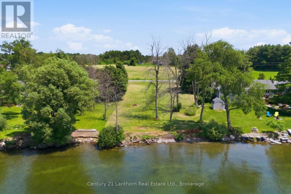 View of a waterfront lot with grass, trees, and rocky shoreline. - Beside 153 Prinyers Cove Crescent, Prince Edward County