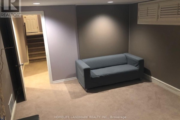 A living room area in the basement with a grey sofa, beige carpet, and a hallway to stairs. - Bsmt - 10 Oscar Court, Toronto