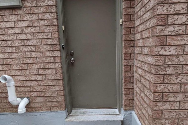 Exterior view of a metal basement door on a brick wall with white drain pipes. - Bsmt - 104 Randall Avenue, Markham